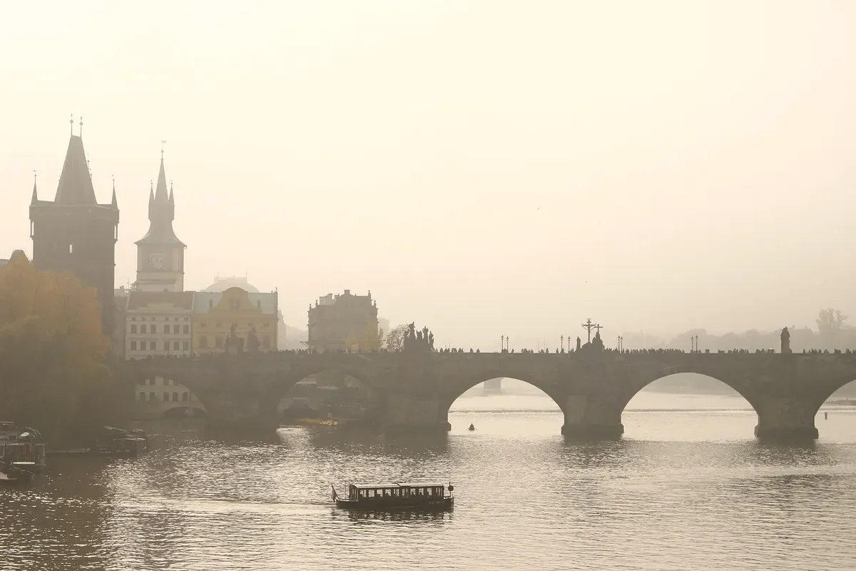 Cầu Charles bắc qua sông Vltava ở Praha trong sương sớm, có hai tháp cầu và một chiếc thuyền nhỏ trên mặt nước